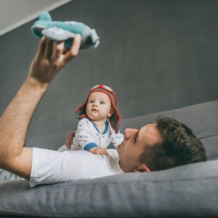 father holding toy plane while lying on sofa and playing with infant child in knitted pilot hat