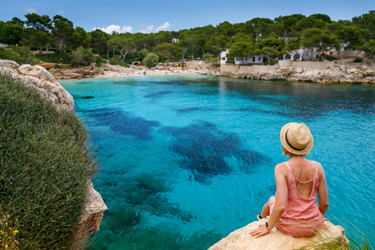 A woman leisurely sitting on a cliff, looking out at Cala Gat beach in Mallorca